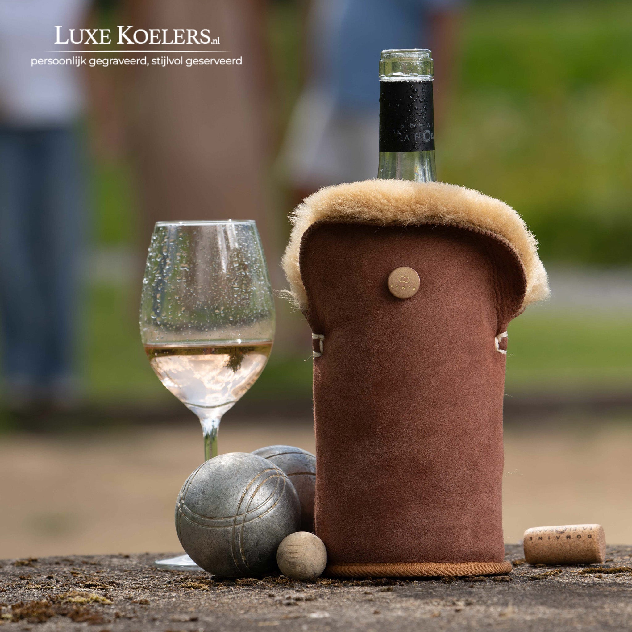 Kywie model Tulip voor wijn en normale champagne flessen.De wijnkoeler is volledig gemaakt van natuurlijke schapenvacht. Koeler met fles en glas met wijn. Naast koeler liggen twee ijzeren jeu de boulle ballen en het cochonnet balletje. Achtergrond vaag mensen buiten