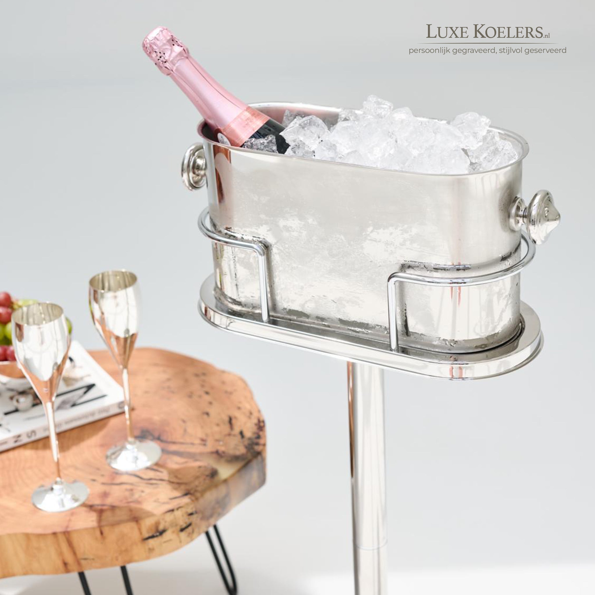 sfeerfoto van koeler met ijs en fles. Daarnaast houten tafel met twee zilverkleurige champagneflutes. Achtergrond licht grijs.
