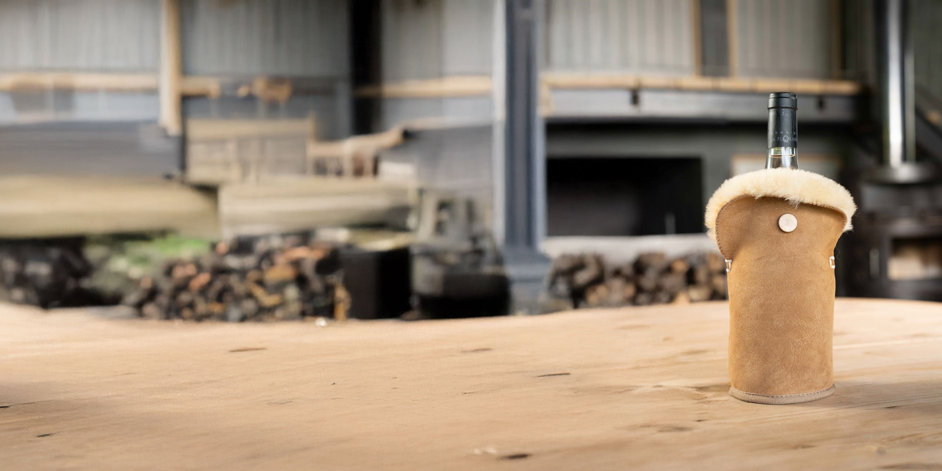 Vachtkoeler van camel kleurige suede met beige schapenvacht. de koeler staat op een houten ondergrond met vaag op de achtergrond een schuur met houten blokken om vuur te stoken