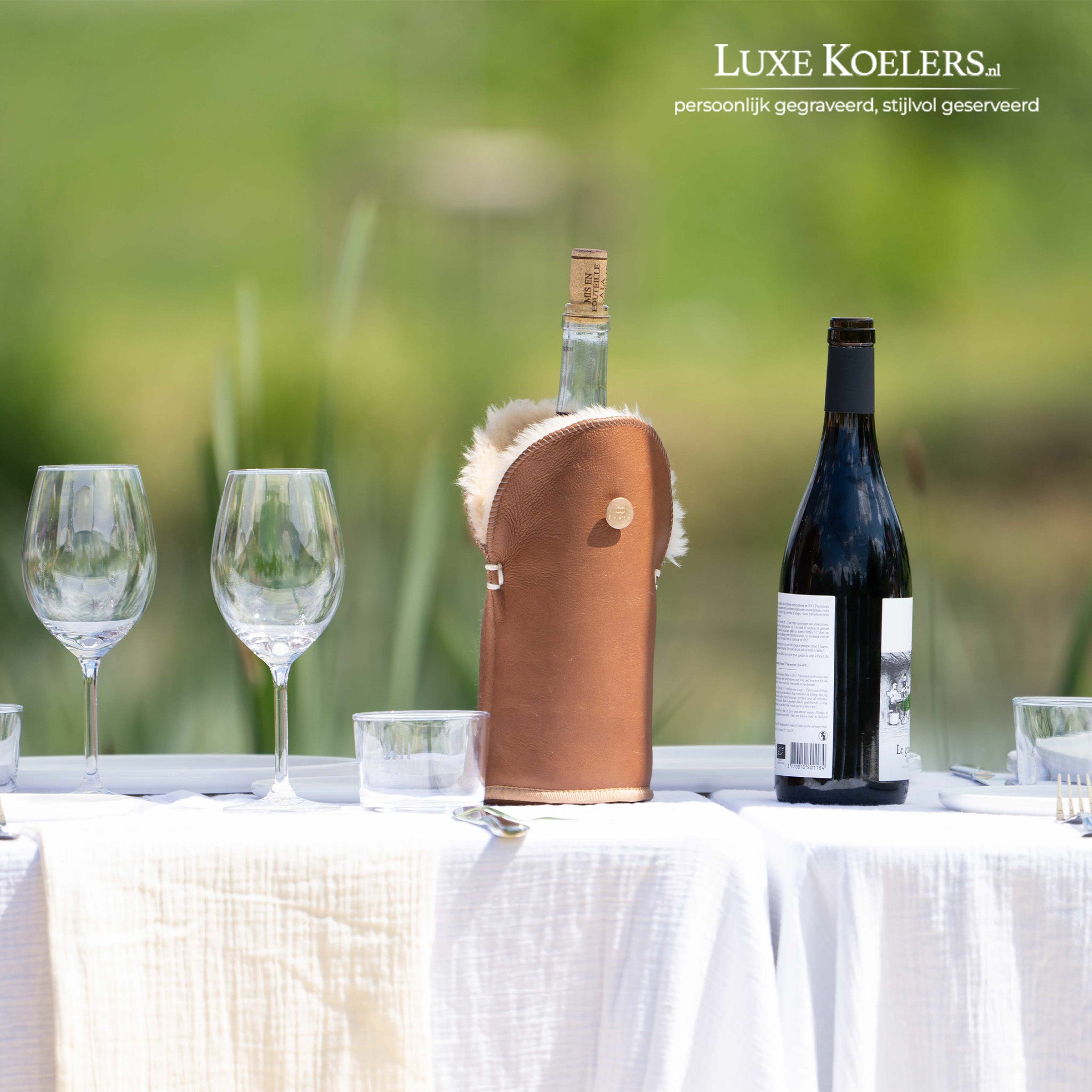 Kywie model Tulip voor wijn en normale champagne flessen.De wijnkoeler is volledig gemaakt van natuurlijke schapenvacht. Nappa koeler camel op tafel in tuin met wit tafelkleed en twee glazen. los naast koeler een fles rode wijn