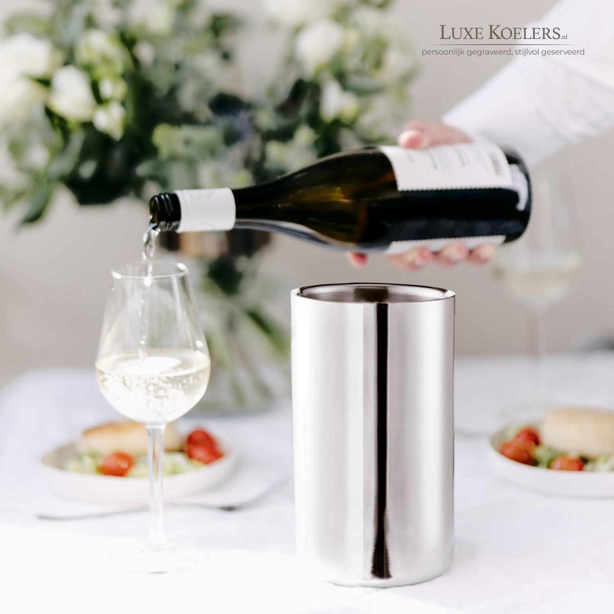 koeler met hand aan fles welke uitschenkt in wijnglas bloemen en eten op achtergrond